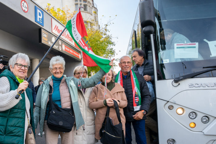 A fotón autóbuszról leszálló idős emberek láthatóak, egyikük kezében magyar nemzeti színű zászló